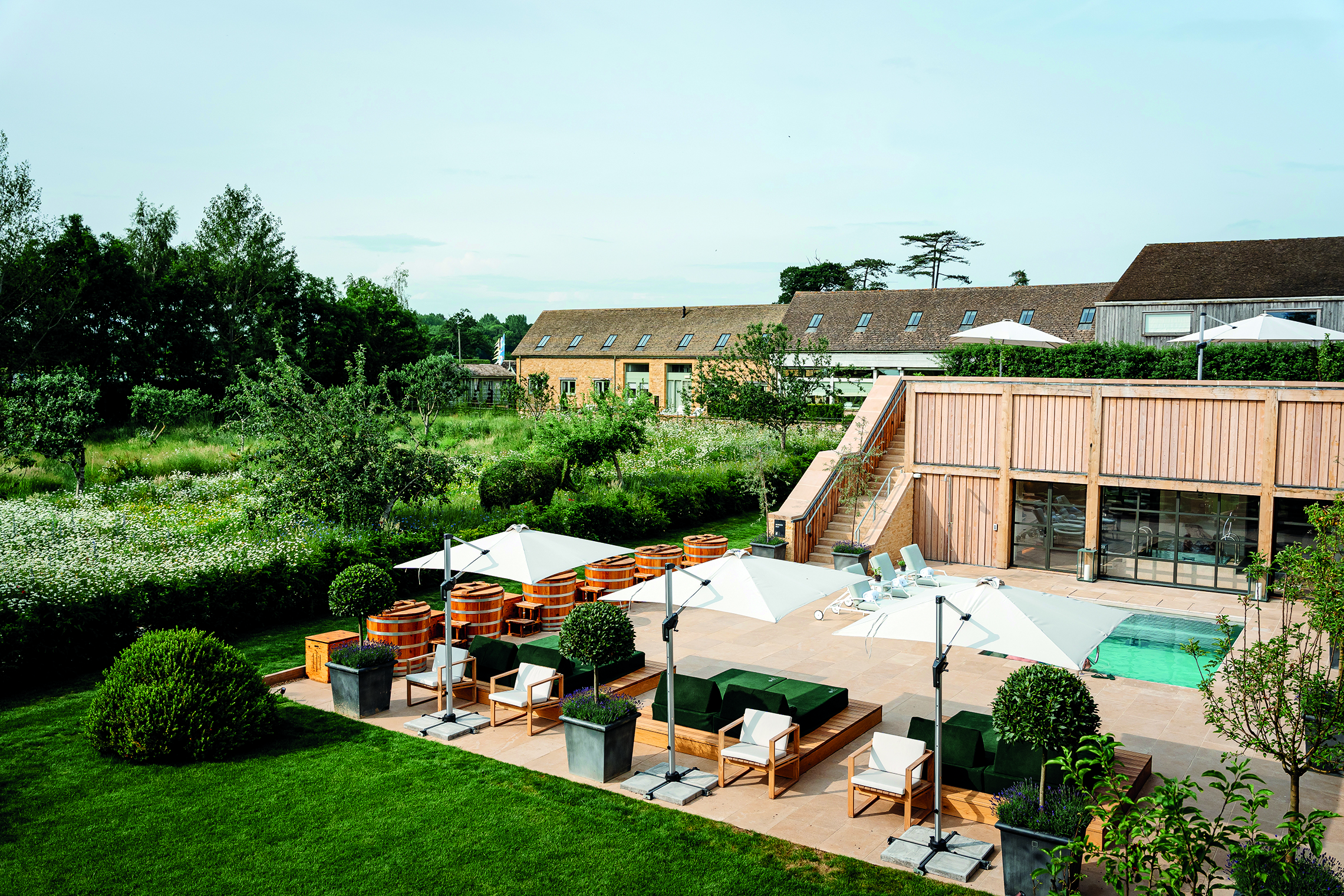 Investment, Tech & Motoring trends 2025 - The Club by Bamford exterior with an outdoor spa area with wooden hot tubs, green lounge chairs, white umbrellas, a pool, and a rustic wooden building surrounded by greenery and wildflowers