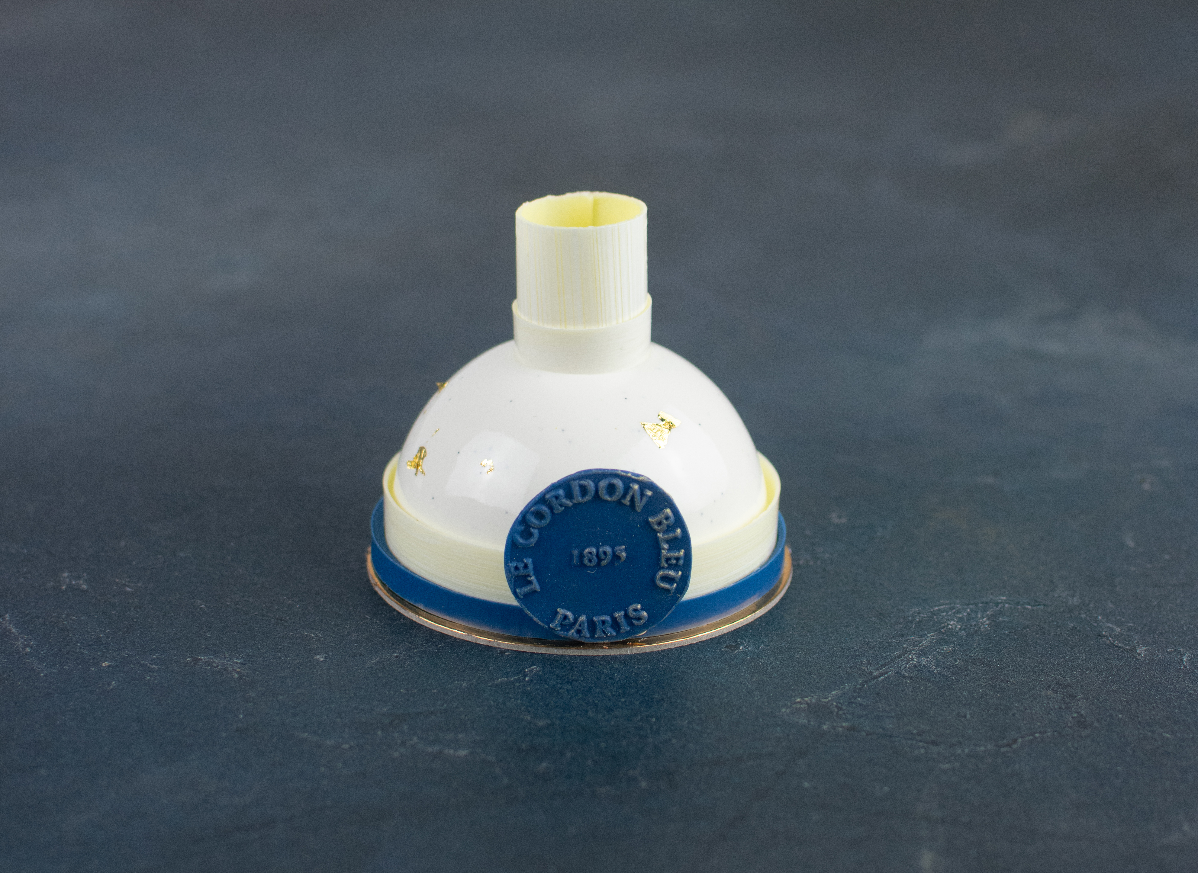 Le Cordon Bleu London - A close-up of a white gateau with a blue base, adorned with the Le Cordon Bleu logo and "Paris" written on it. The bell features a subtle golden decoration and is placed on a dark surface, emphasizing its detailed craftsmanship.