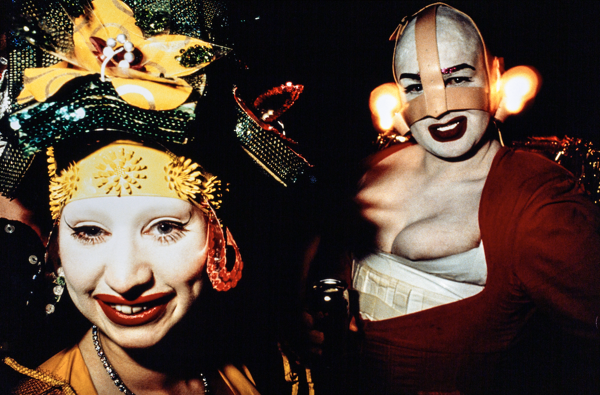 Leigh Bowery Tate Modern - A close-up of two individuals in dramatic, colorful costumes and makeup. One wears a yellow headpiece with glittering accents, while the other sports a red and white costume with exaggerated makeup, contributing to their avant-garde appearance