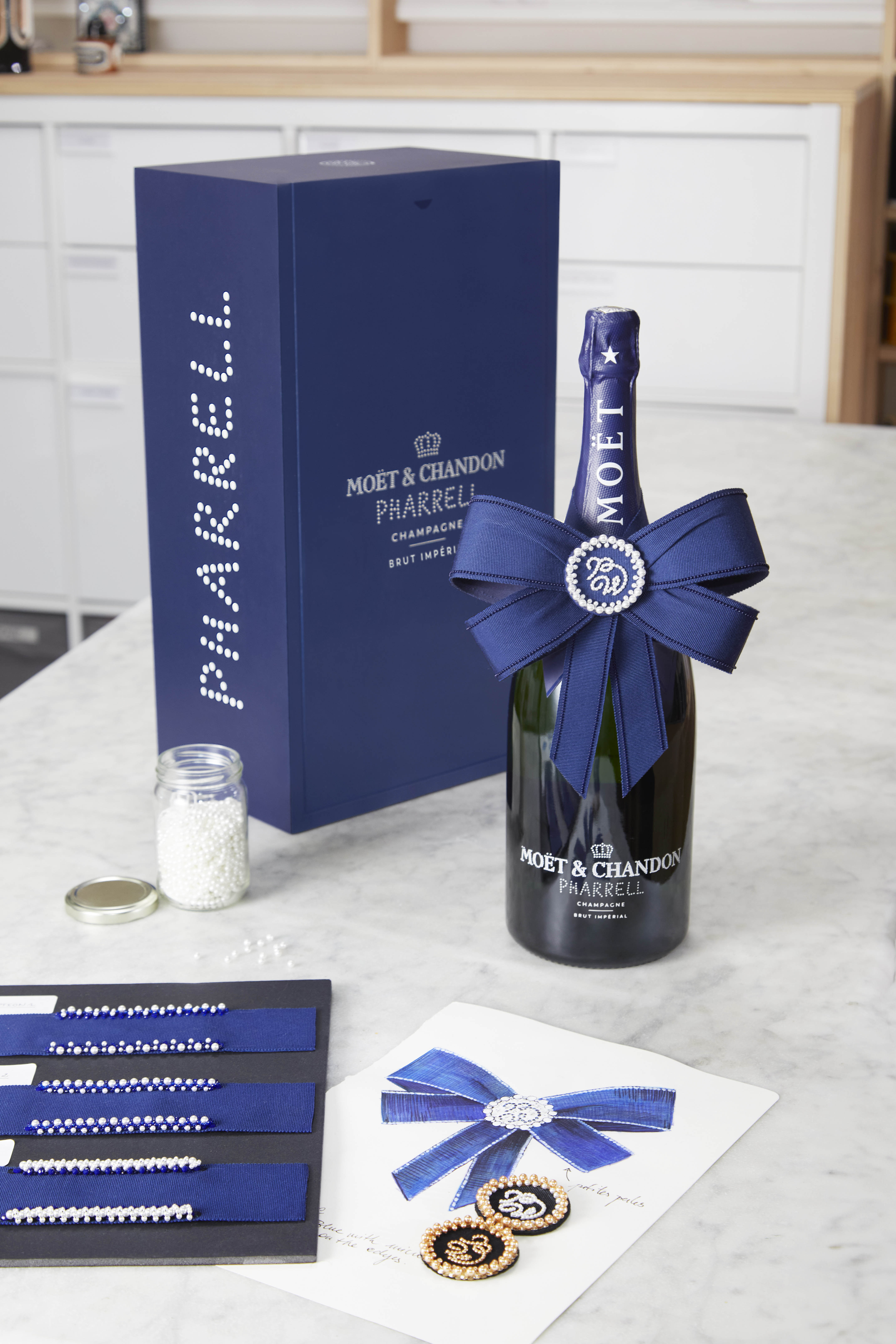 Pharrell Williams Moet Chandon - The image shows a bottle of Moët & Chandon Pharrell Williams limited edition champagne adorned with a navy blue satin bow, placed beside its matching gift box. The setup includes a jar of pearl beads, a design sheet with an illustration of the bow, and a set of pearl-embellished straps, emphasising the luxury and craftsmanship behind the collaboration.