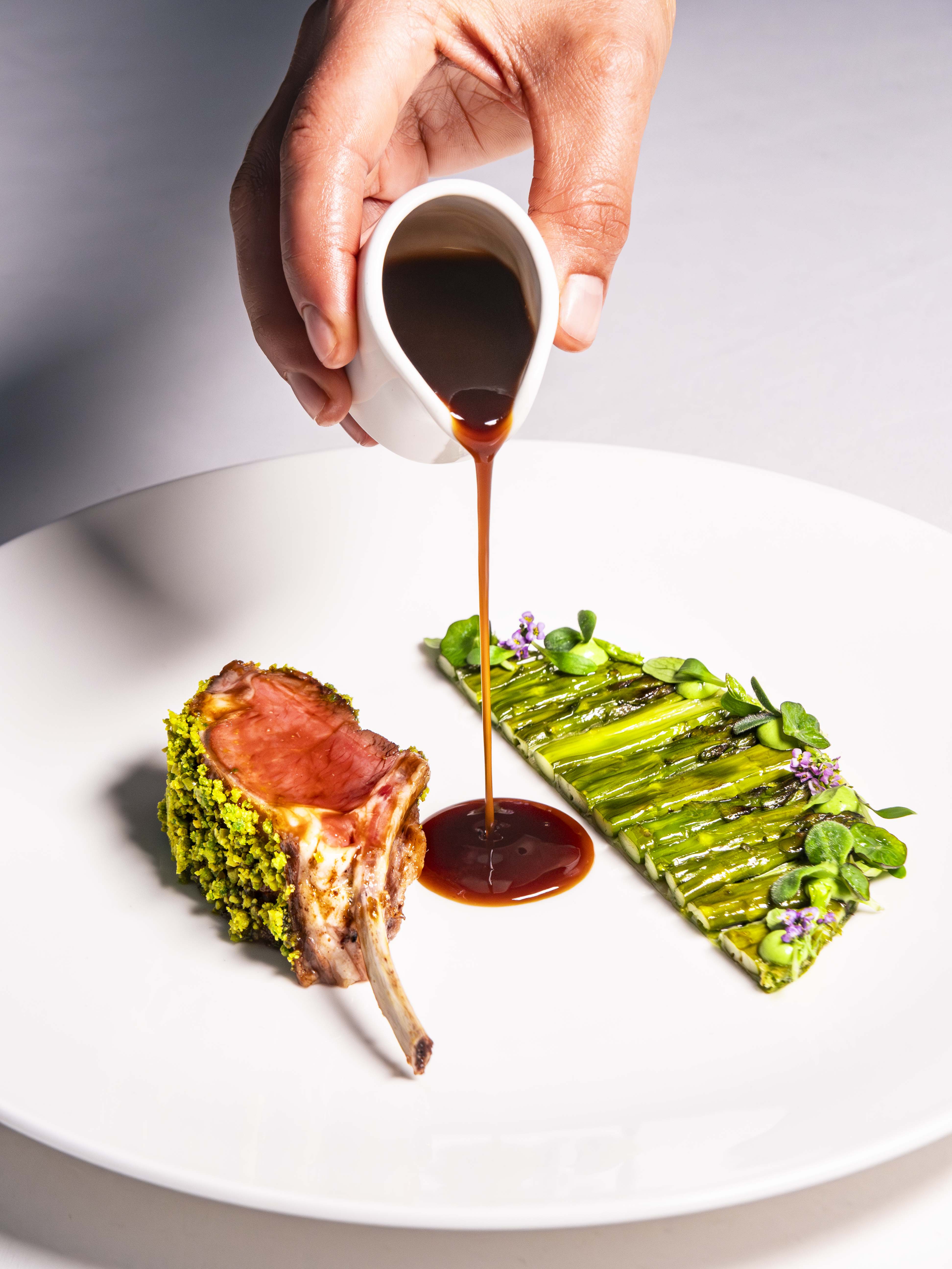 Le Cordon Bleu London - A close-up of a hand holding a small white gravy boat, pouring a rich brown sauce onto a beautifully plated dish with a pistachio-crusted lamb chop and grilled asparagus arranged artistically with herbs.