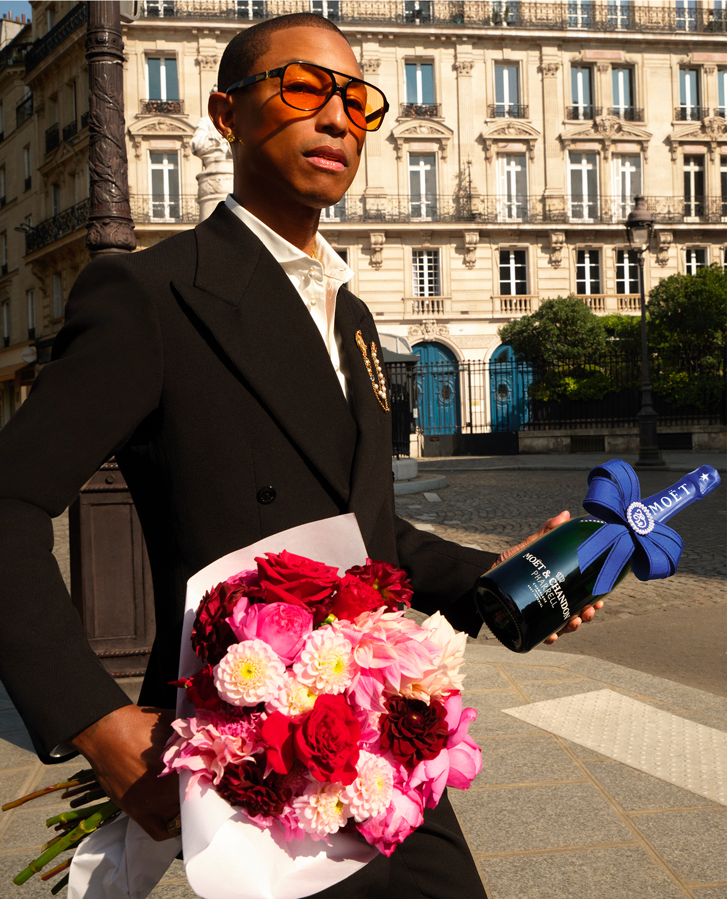 Pharrell Williams Moët & Chandon - Pharrell williams wearing a suit and walking in front of creamy white building holding a bouquet of pink and red flowers and a bottle of blue champagne