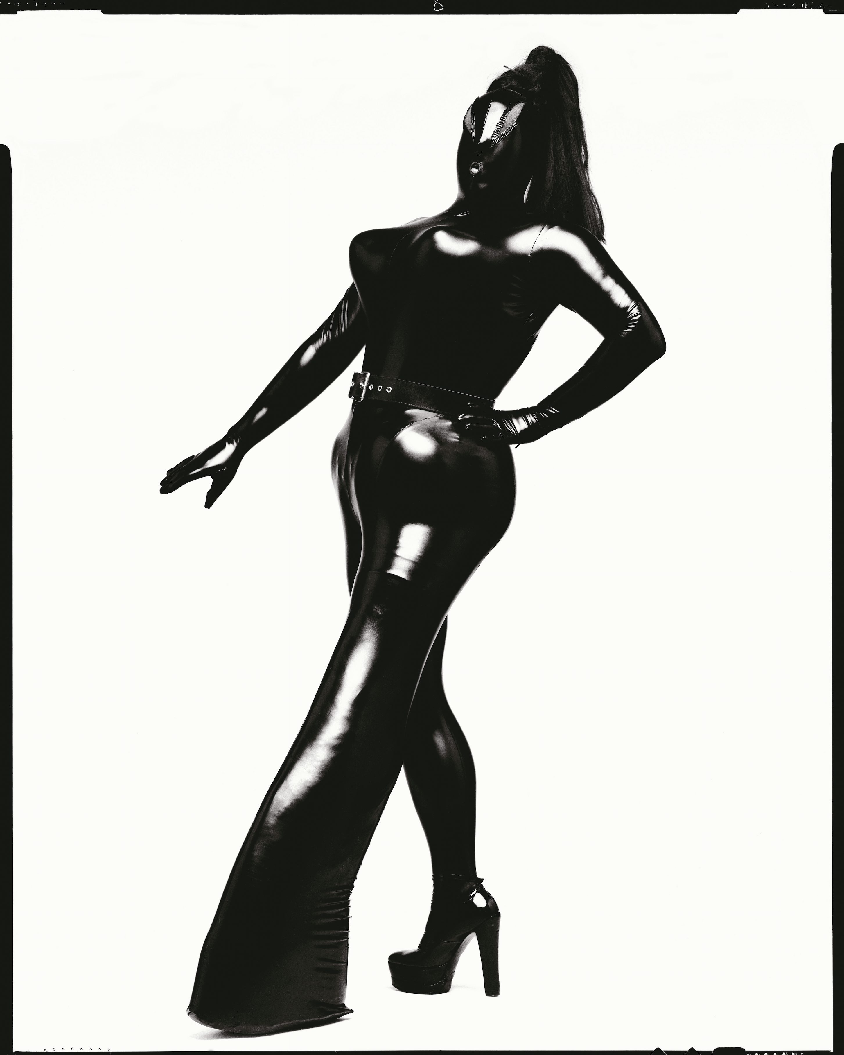 Leigh Bowery Tate Modern - A dramatic black-and-white photograph of Leigh Bowery striking a pose in a full-body, shiny black latex outfit, covering the body, including the head. The outfit features a tight fit with a long, flowing leg, a high heel, and a belt accentuating the waist. The model's silhouette stands out against a stark white background, creating a striking and futuristic appearance