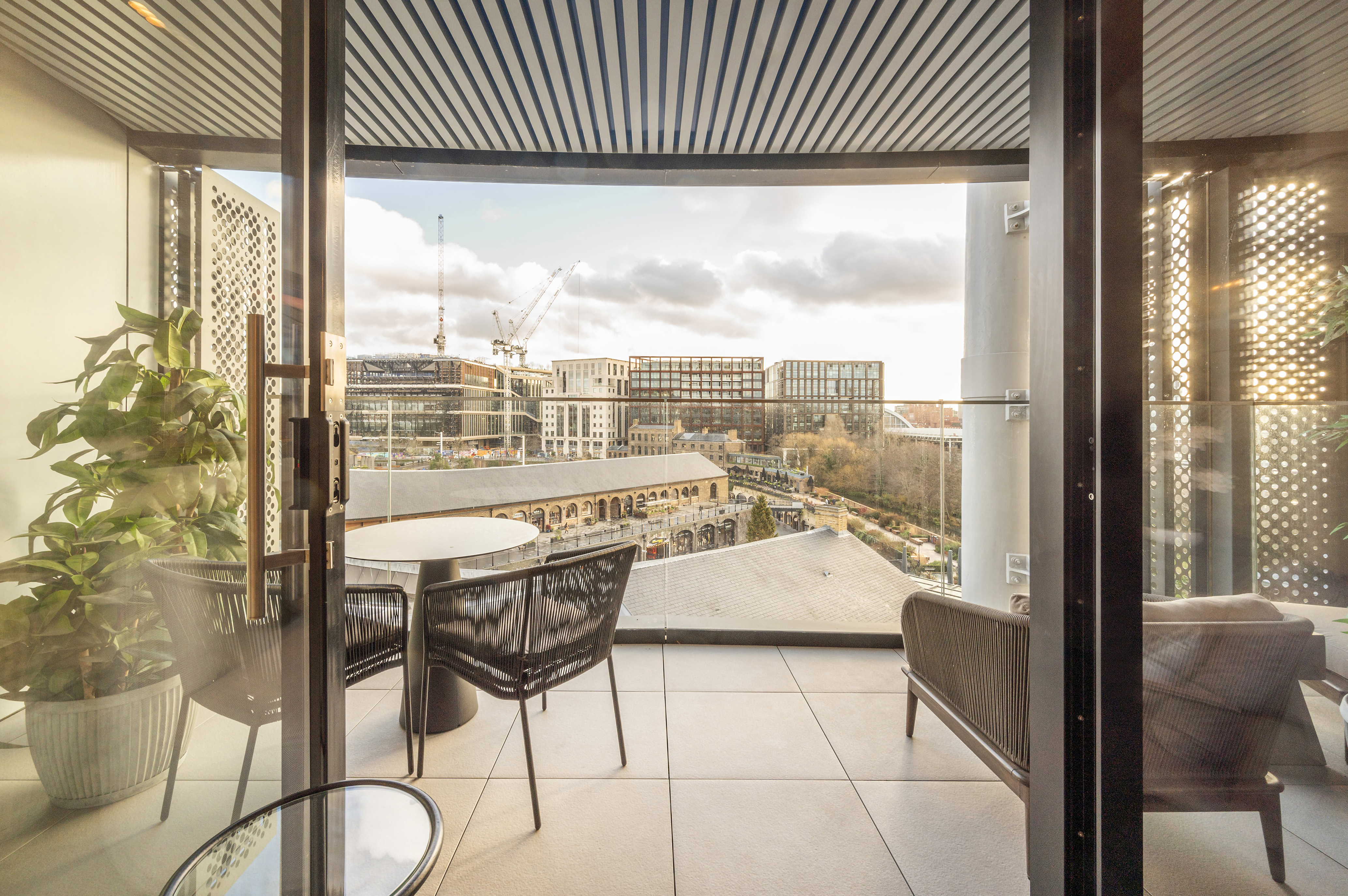 Emerging London Boroughs New Luxury Property - A modern apartment balcony with a glass railing, overlooking a cityscape with construction cranes, office buildings, and a historic market-style structure. The balcony features a round dining table with two chairs, a lounge chair, and a potted plant