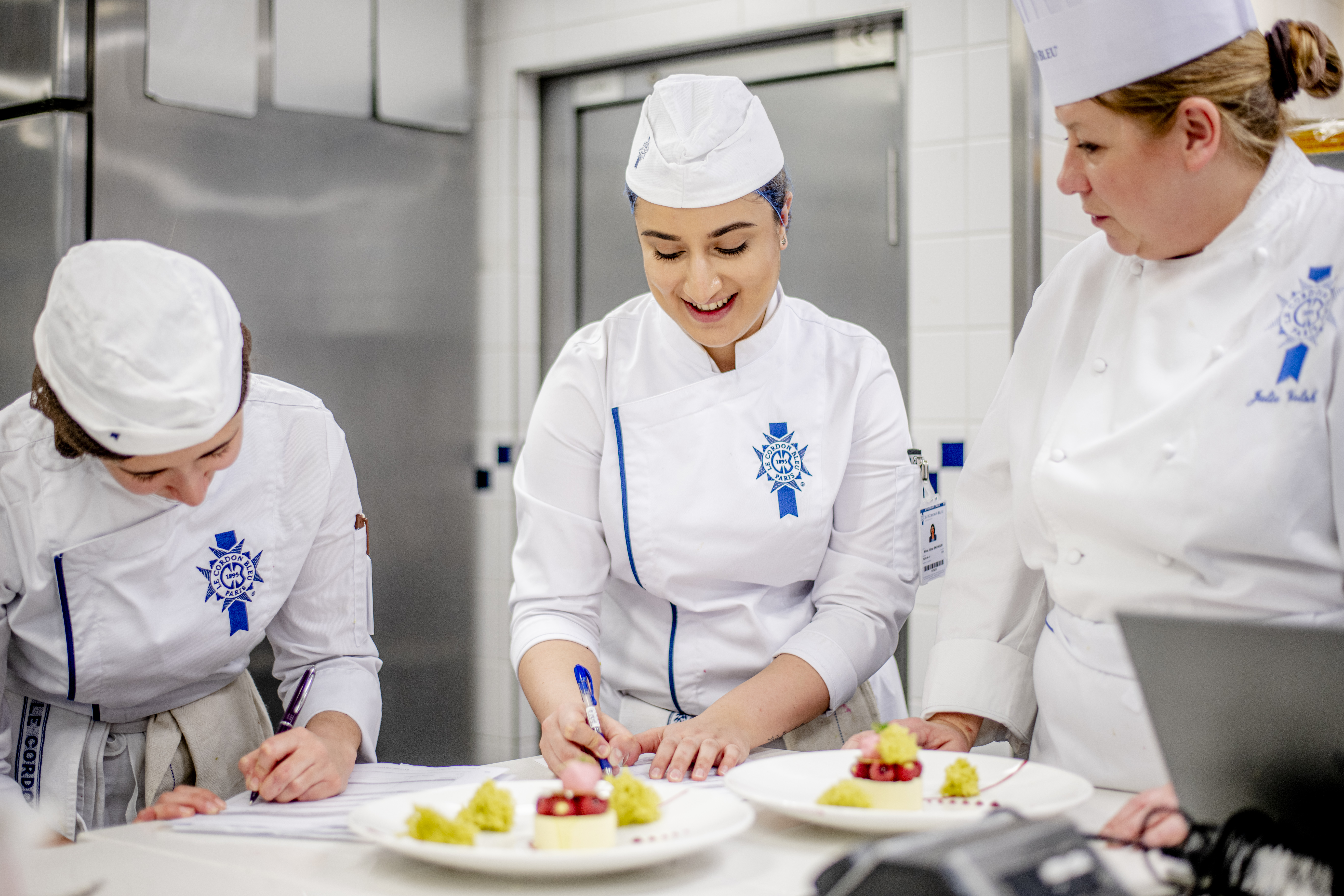 Le Cordon Bleu London - A group of culinary students in white Le Cordon Bleu uniforms work together in a kitchen. One chef is smiling while writing, while another arranges a dessert with fresh berries and cream on a white plate. The kitchen background is filled with stainless steel equipment.