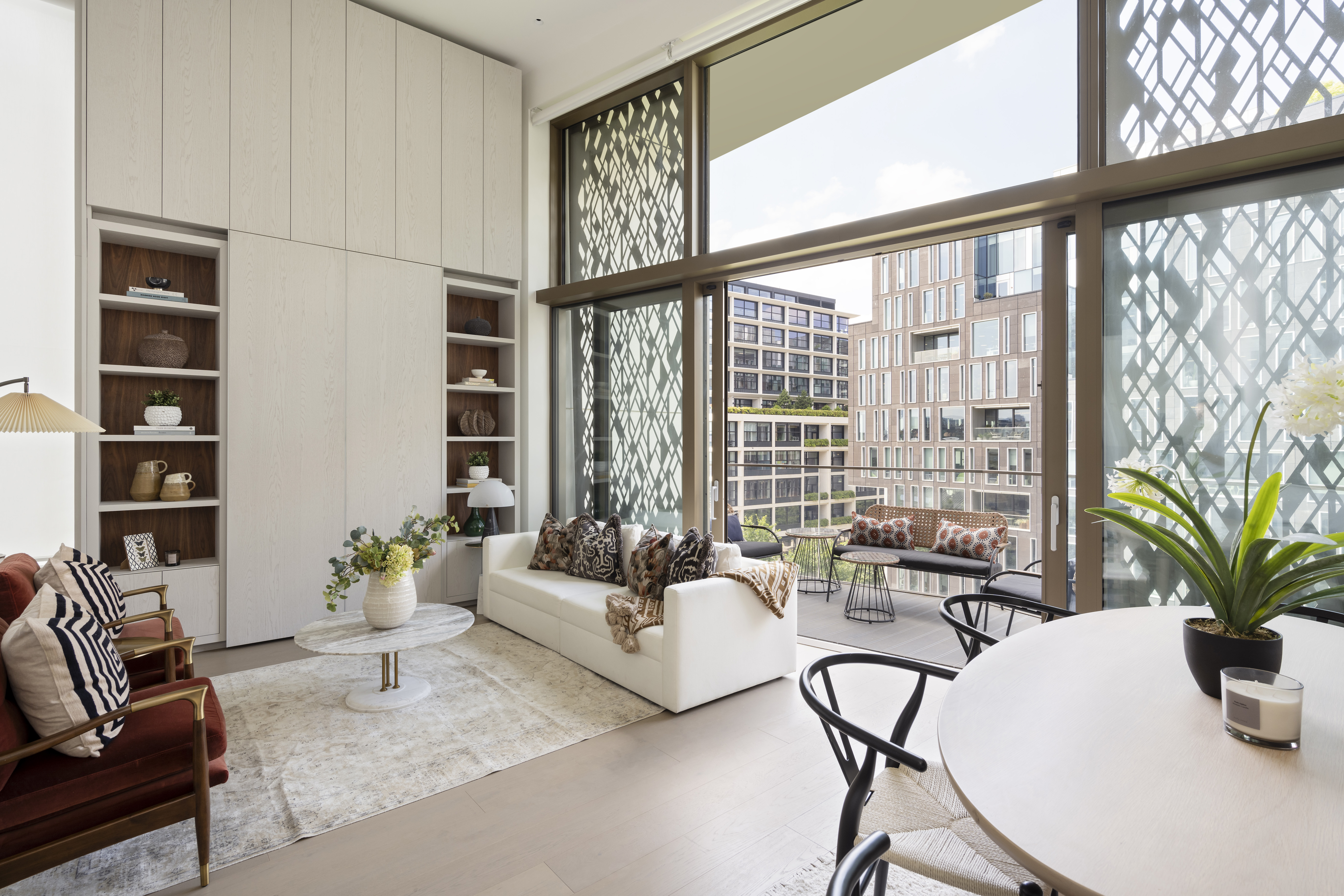 Emerging London Boroughs New Luxury Property - A stylish open-plan living room in a luxury apartment with floor-to-ceiling windows leading to a spacious balcony. The room features a white sofa, built-in wooden shelves, and a round dining table with black chairs. The interior design blends neutral tones with warm textures, and the large windows allow natural light to flood the space.