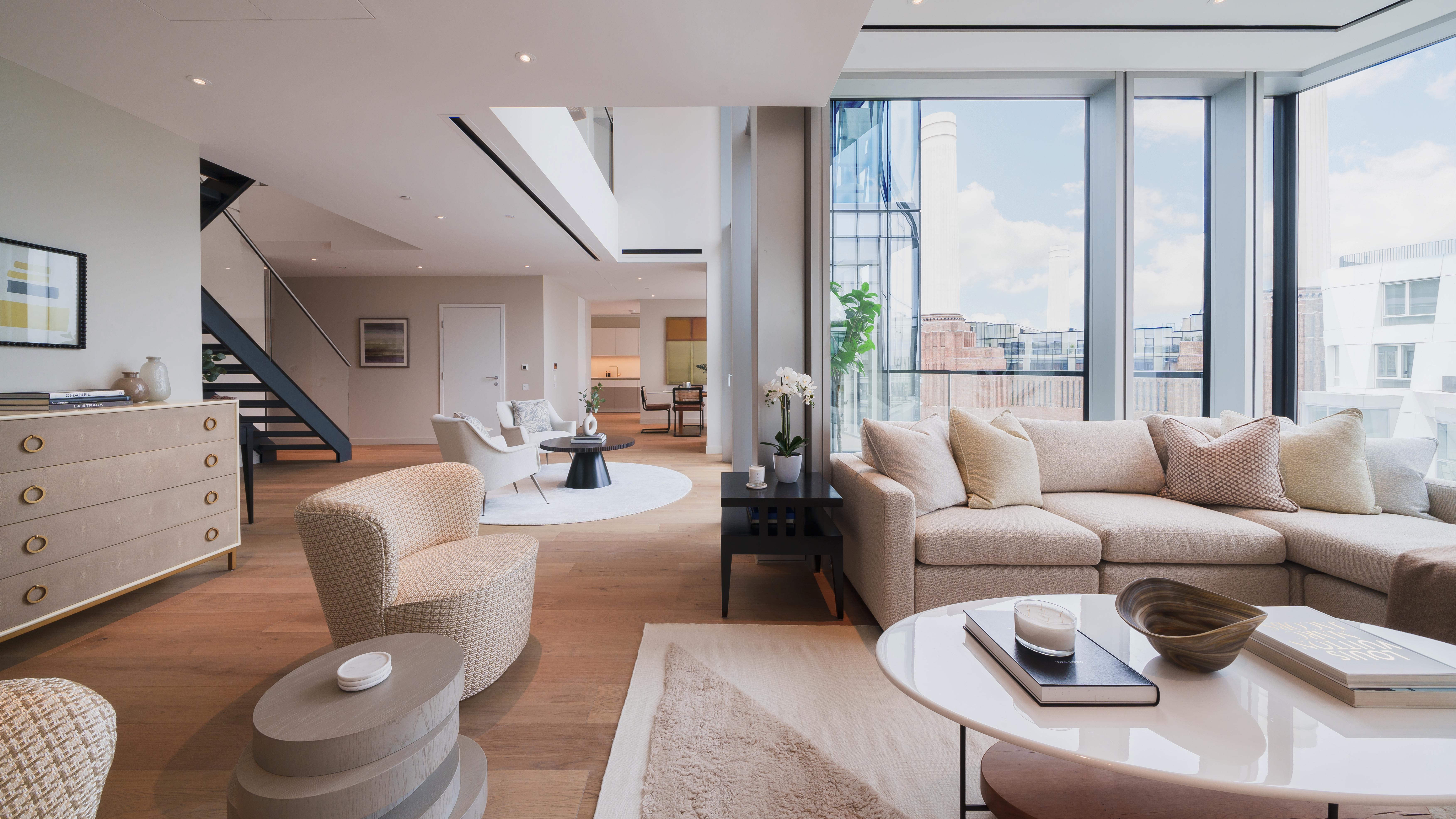 Emerging London Boroughs New Luxury Property - A modern and stylish living room featuring a light beige sofa with textured pillows, a round coffee table with decorative items, and a small dark side table with books and a plant. The room has large windows providing a view of a nearby building and a blue sky. A staircase with a sleek design leads to the upper level, and light wood flooring adds warmth to the space.