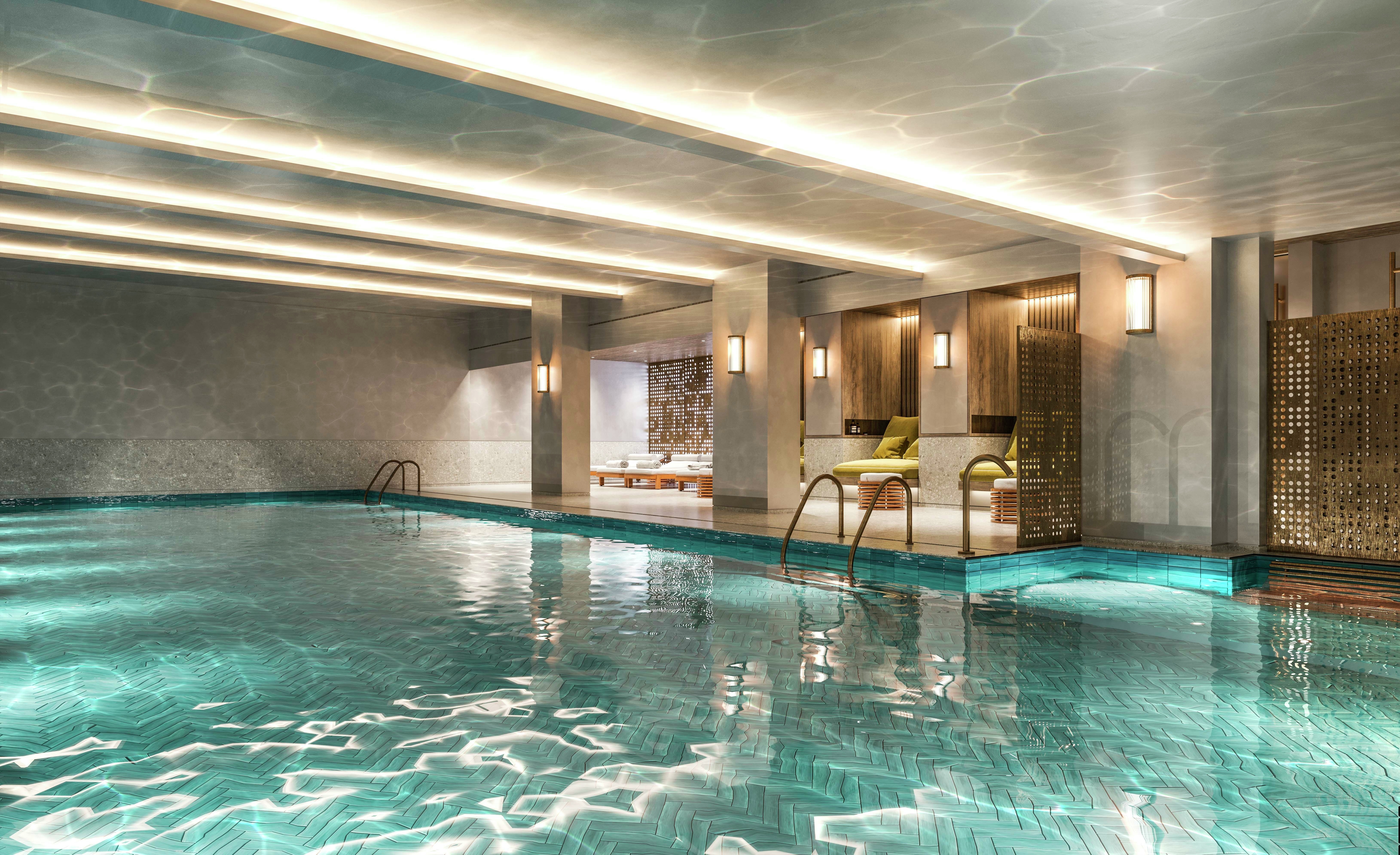 Emerging London Boroughs New Luxury Property - A luxurious indoor swimming pool area with blue-tiled flooring, illuminated by soft lighting. The pool has clear water with a reflective surface, and a set of stairs leading into the pool. A relaxing lounge area with modern furniture can be seen in the background.