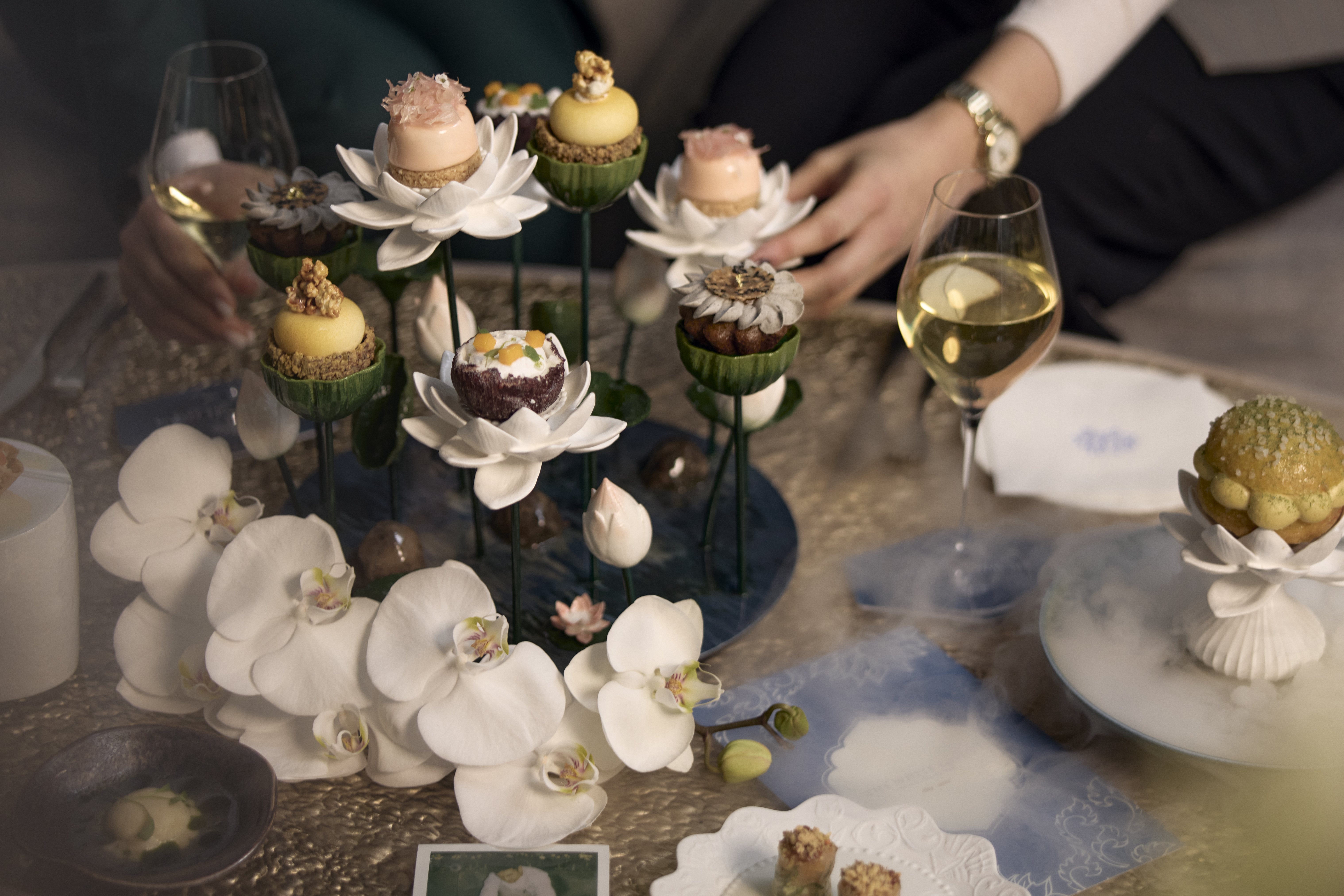 White Lotus London -  A beautifully arranged afternoon tea setup featuring delicate pastries presented on tall, elegant flower-shaped stands. The treats are decorated with intricate toppings like flowers, nuts, and fruit, surrounded by white orchid flowers. A glass of wine is in the background, and the table has a luxurious gold-coloured surface with fine porcelain plates and napkins.