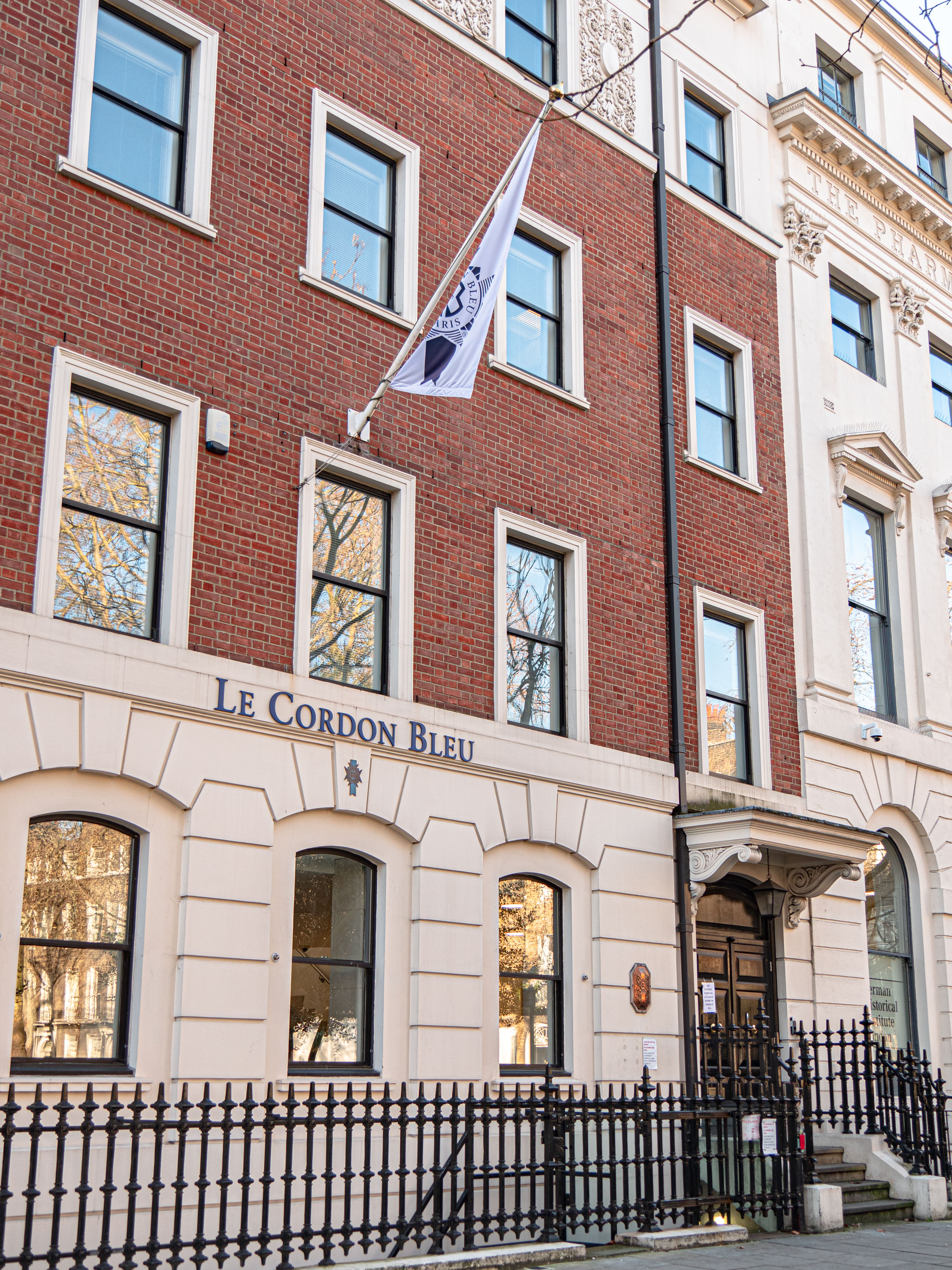 Le Cordon Bleu London - A brick building with large windows and a white flag featuring the Le Cordon Bleu logo. The flag is fluttering in front of the building, which is located in a tree-lined street.