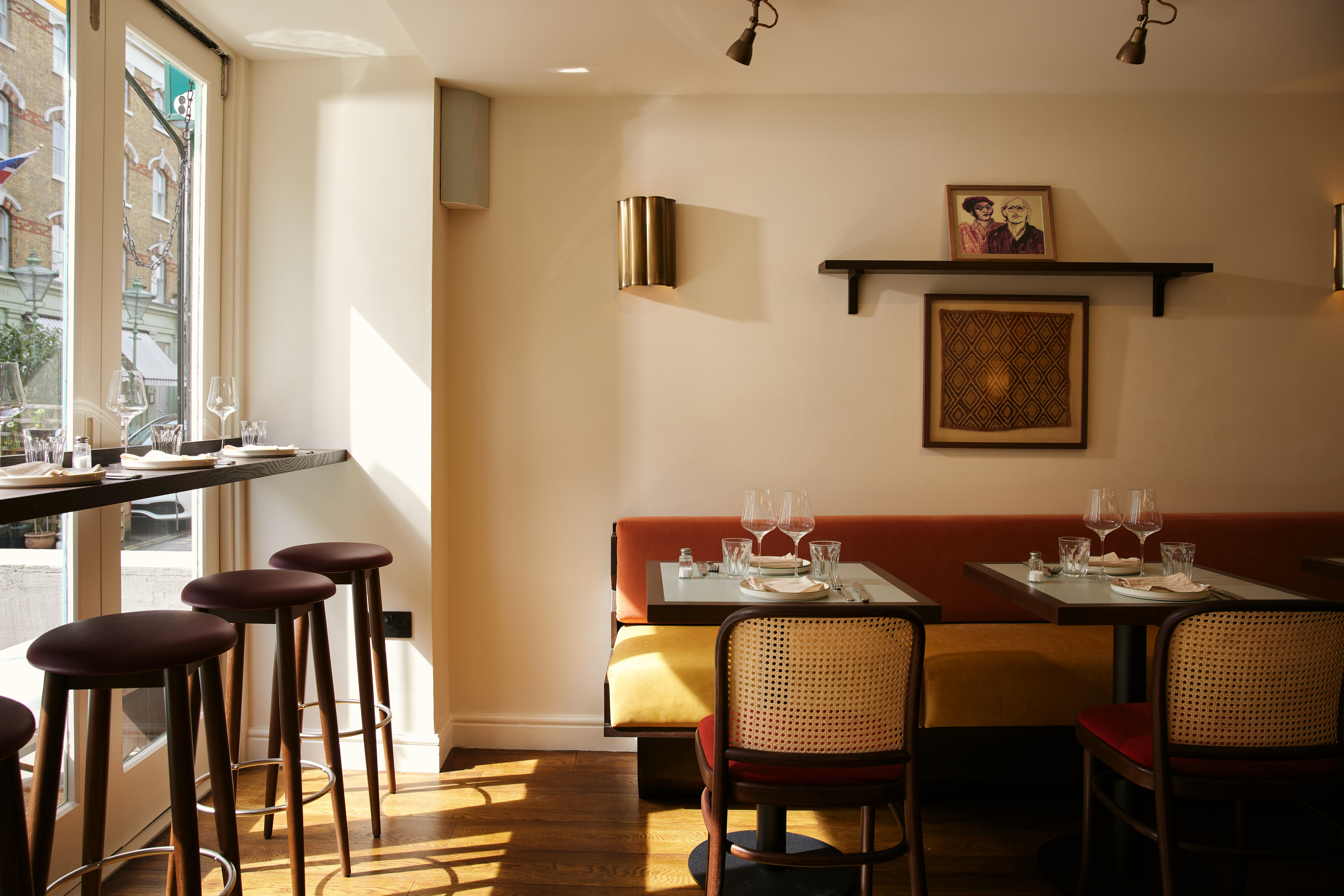 Art restaurants in London - The restaurant interior of July in Fitzrovia with a built-in bench with mustard upholstery, laid tables, cane-backed chairs, and a high-top bar with wooden stools.