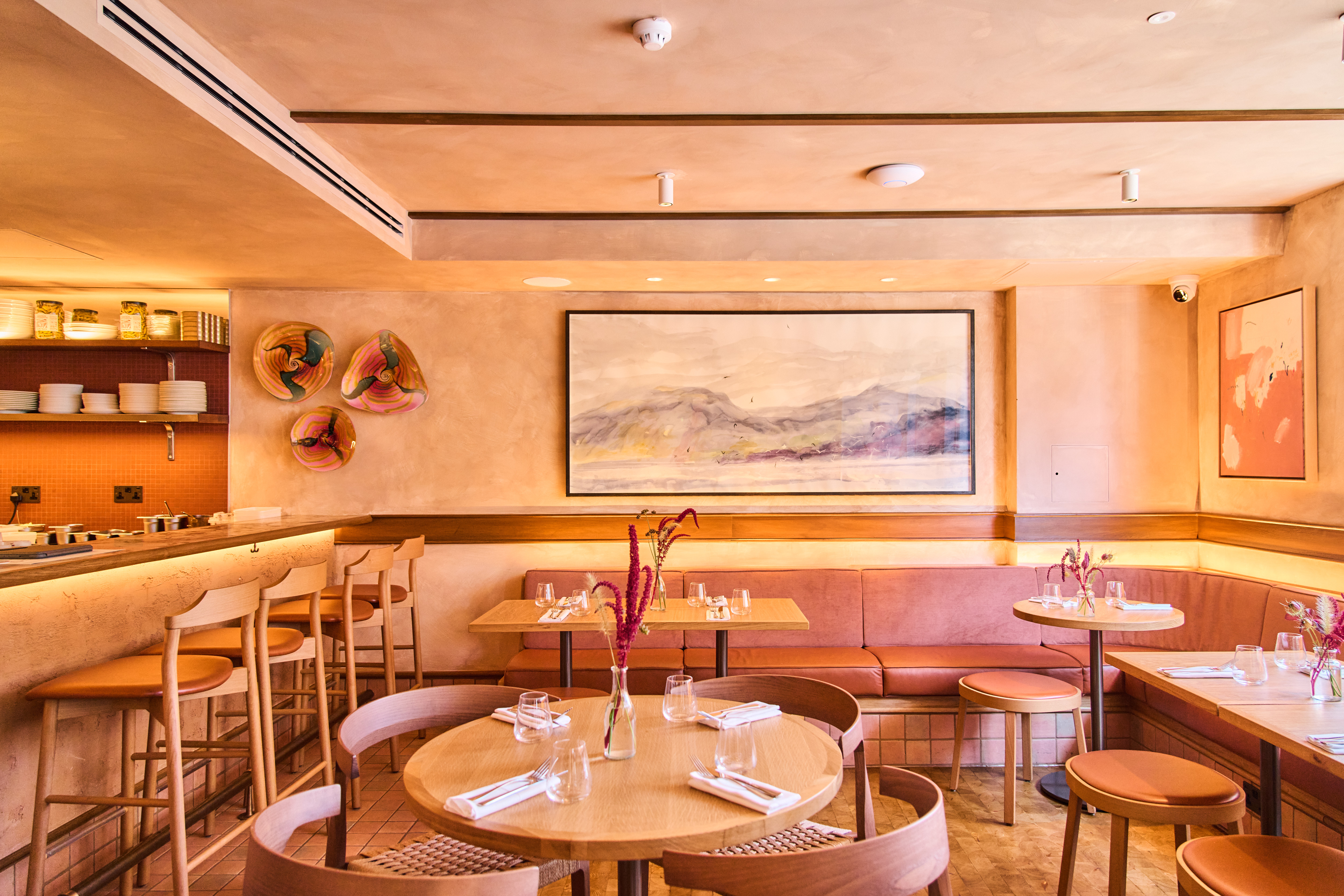 Art restaurants in London - The restaurant interior of Lolo on Bermondsey Street with wooden furniture, and a long cushioned bench alongside laid tables. Artwork and decorative ceramic pieces can be seen on the walls.