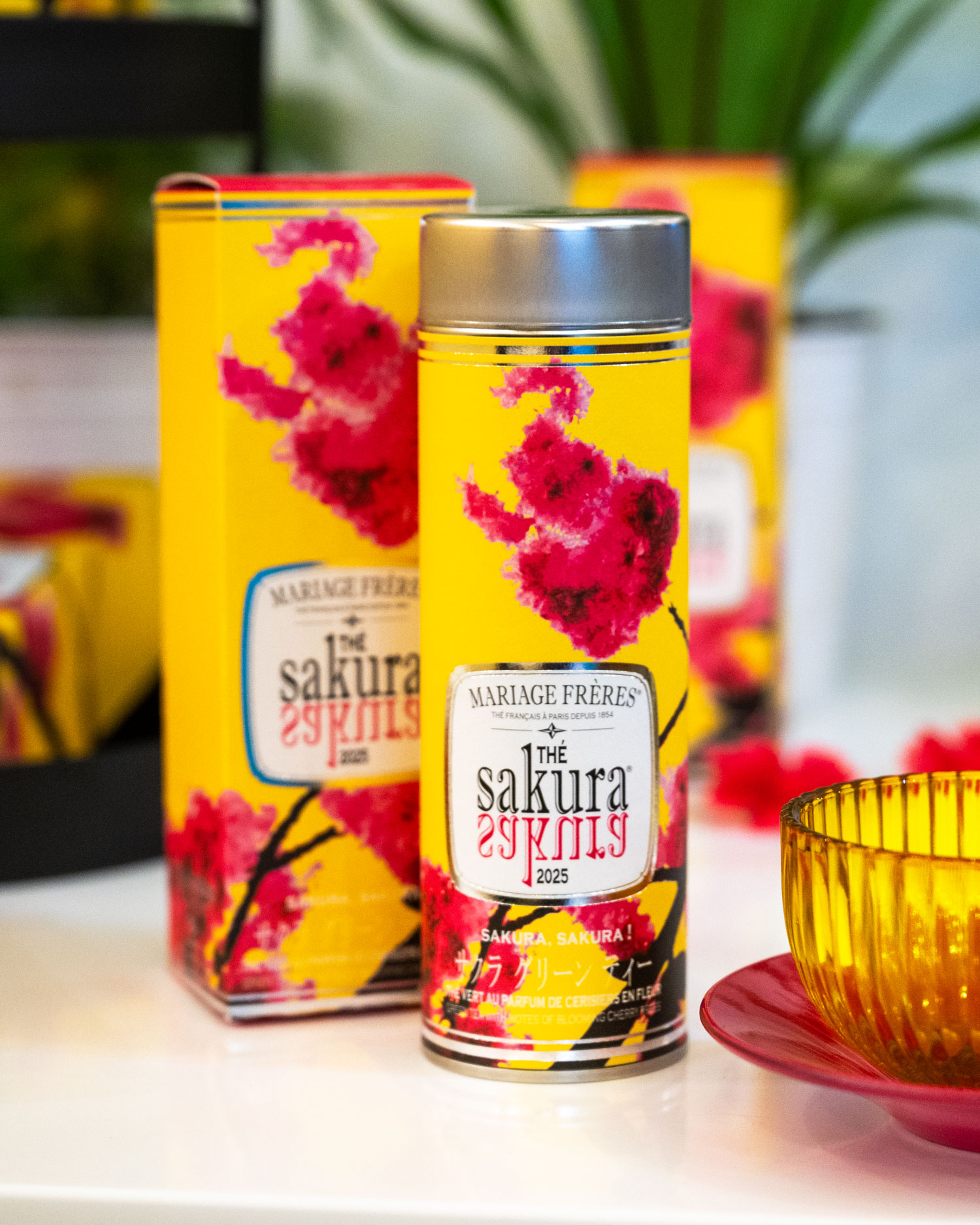 Mariage Frères Japanese floral teas spring - The image features a vibrant yellow tea canister from Mariage Frères, decorated with pink sakura flowers. The packaging, labeled "The Sakura 2025," highlights the floral theme with a design that combines the colors pink, yellow, and red. A matching yellow bowl and red plate are placed in the foreground, adding to the elegant and colorful presentation. The background is softly blurred with additional canisters visible.