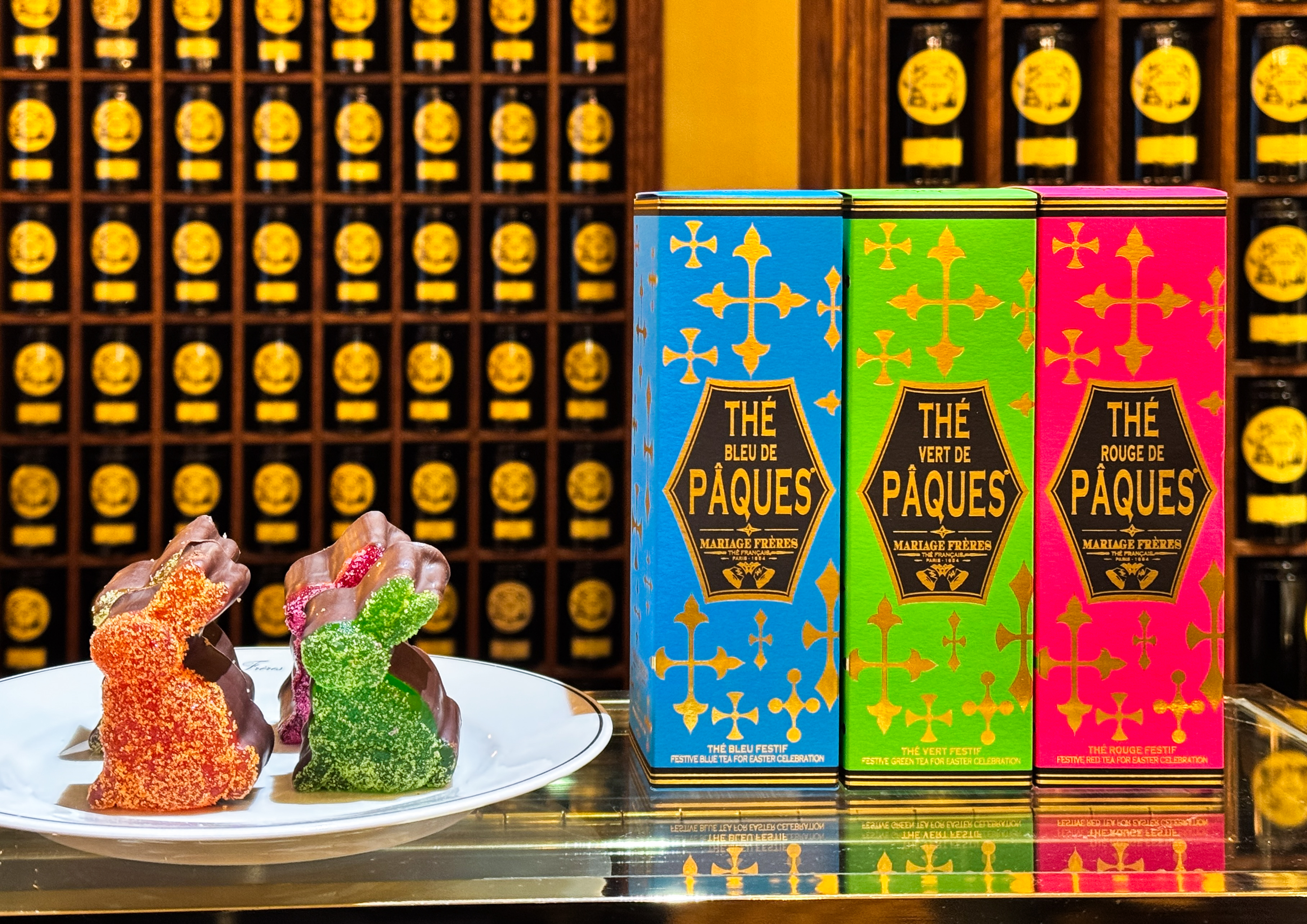 Mariage Frères Japanese floral teas spring - Colourful chocolate Easter bunnies covered in sugar crystals, alongside three vibrant tea boxes labeled Thé Bleu de Pâques, Thé Vert de Pâques, and Thé Rouge de Pâques, displayed in front of a background of Mariage Frères tea tins.