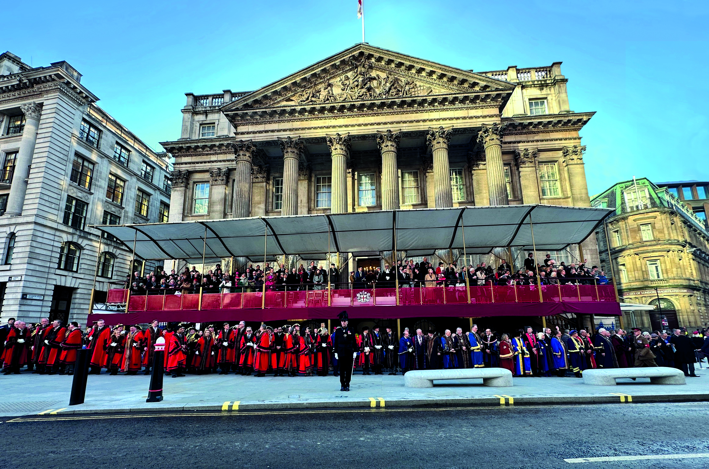 Inside Mansion House Lord Mayor Residence - A large crowd of people, including several individuals in red ceremonial robes, stands on the street in front of a grand classical-style building. The scene is apart of the Lord Mayor's show, with the people gathered under a canopy.