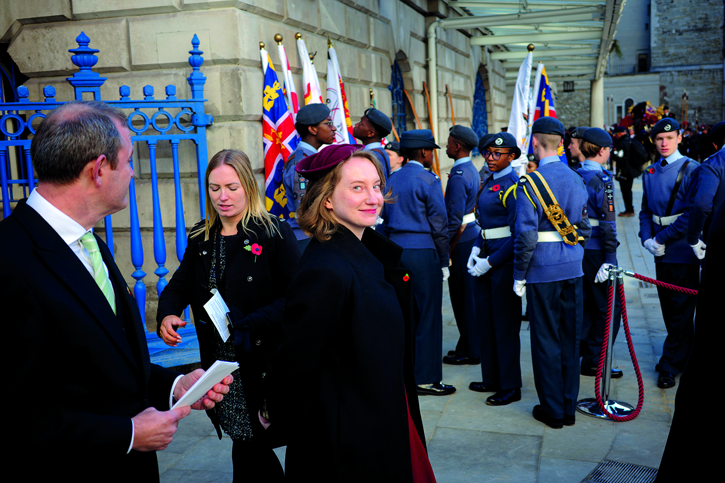 Inside Mansion House Lord Mayor Residence - Caroline Jack, along with a man and another woman, is seen walking outside near a large building with blue gates. The group is dressed formally, and a ceremonial event involving guards in uniforms is taking place in the background.