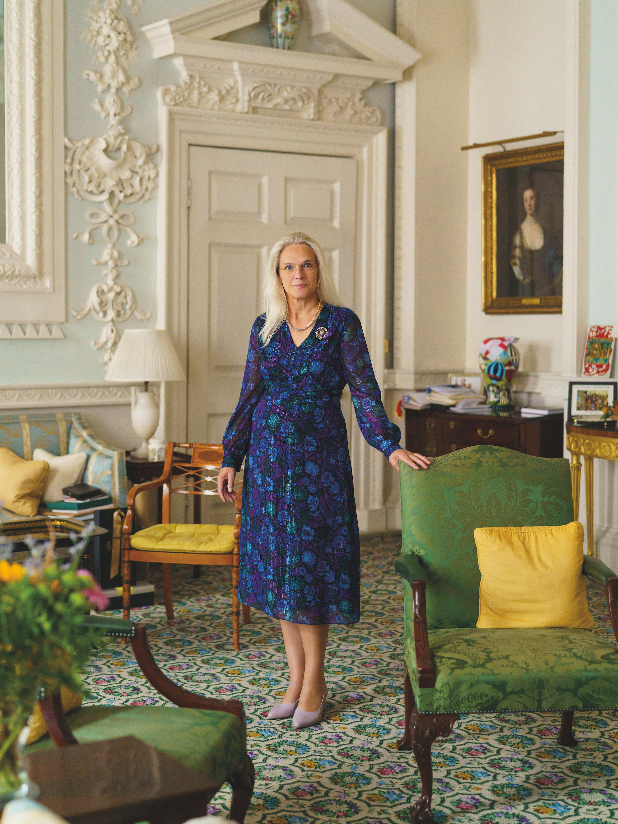 Inside Mansion House Lord Mayor Residence - A Elisabeth Mainelli stands in a formal room, dressed in a blue patterned dress. She is positioned next to a green upholstered armchair, with intricate white detailing on the walls and a portrait visible in the background.