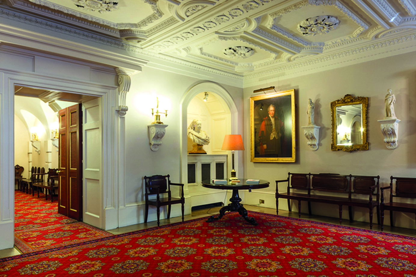 Inside Mansion House Lord Mayor Residence - A traditional, elegant hallway with ornate white walls and a richly patterned red carpet. Several pieces of fine art hang on the walls, and classic wooden benches line the hall. Decorative sculptures are placed along the walls.