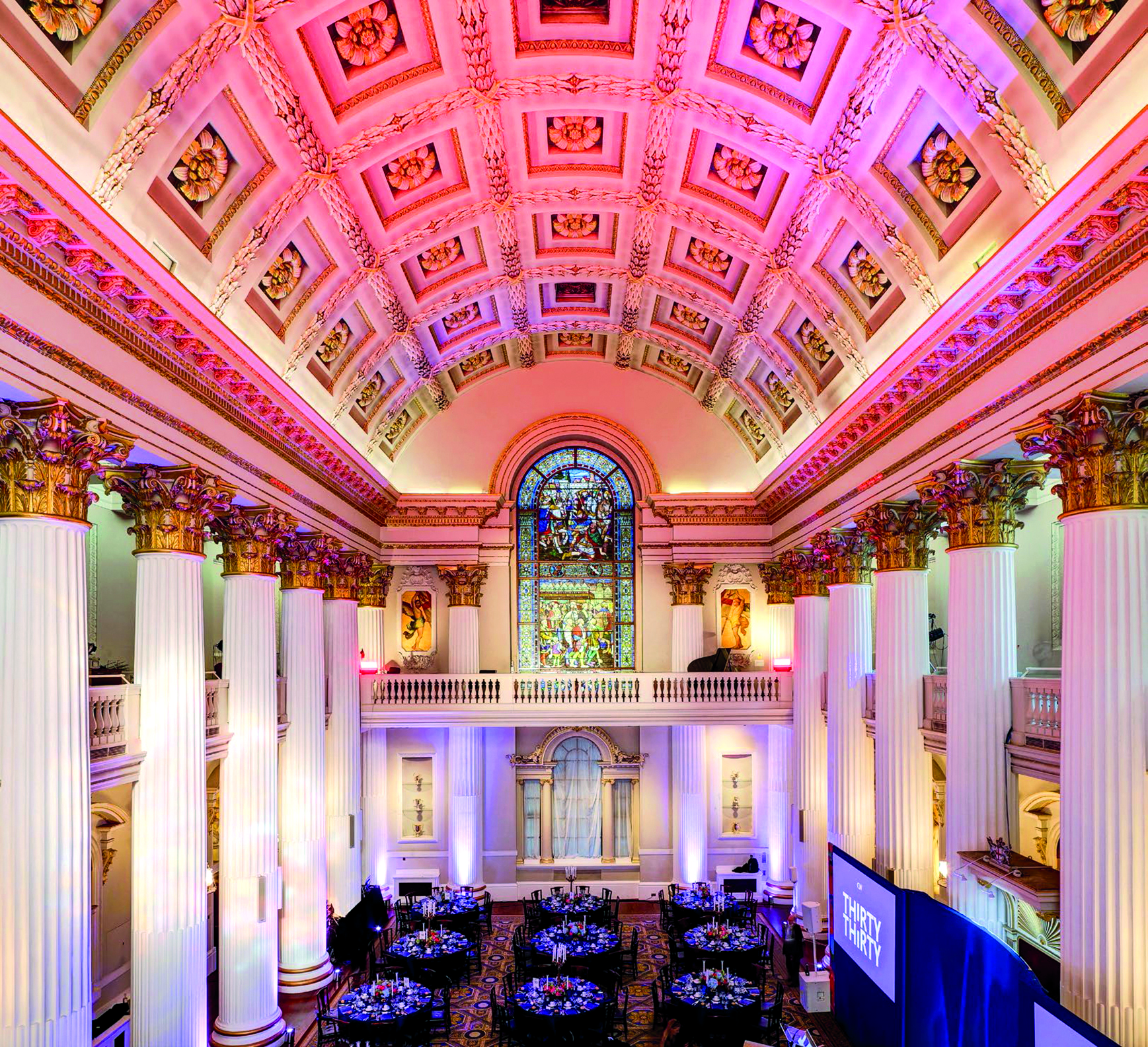 Inside Mansion House Lord Mayor Residence - A wide shot of a grand hall with towering columns, large stained glass windows, and several round tables set for an event. The ceiling is richly decorated with intricate patterns, and the lighting gives the room a vibrant, upscale atmosphere