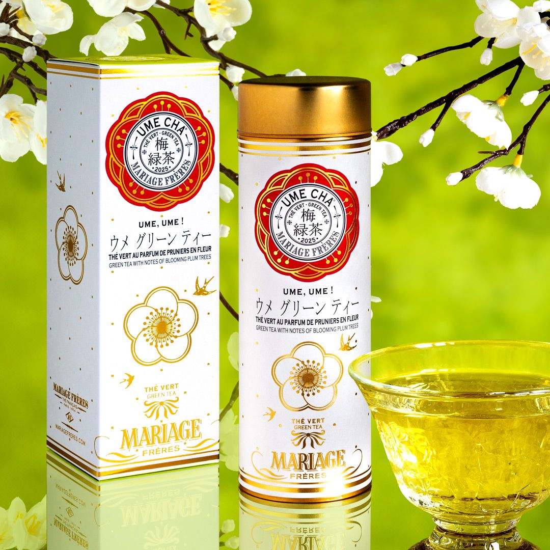 Mariage Frères Japanese floral teas spring - The image features a canister of Mariage Frères "Ume Cha 2025," a green tea infused with the fragrance of blooming plum trees. The canister is white with red and gold accents, adorned with a floral design. In the foreground, there is a delicate glass cup filled with pale yellow tea. The background showcases soft, blurred plum blossoms against a light green backdrop, emphasising the floral and fresh theme.