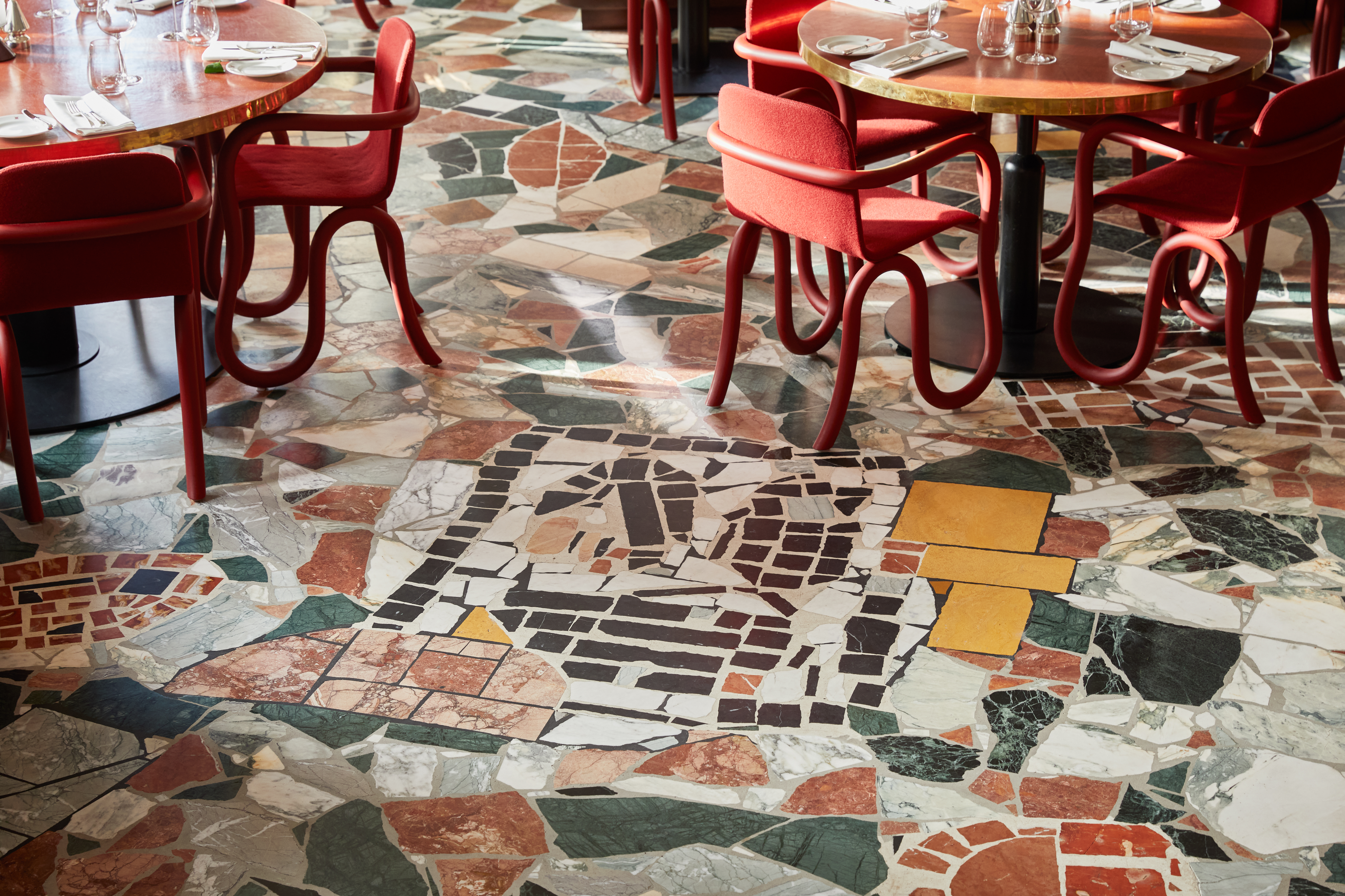 Art restaurants in London - A detailed view of the mosaic floor at Mount St. Restaurant, designed by Rashid Johnson. The floor features an intricate mix of multicoloured marble and stone pieces. Tables with red chairs can be seen to the side.