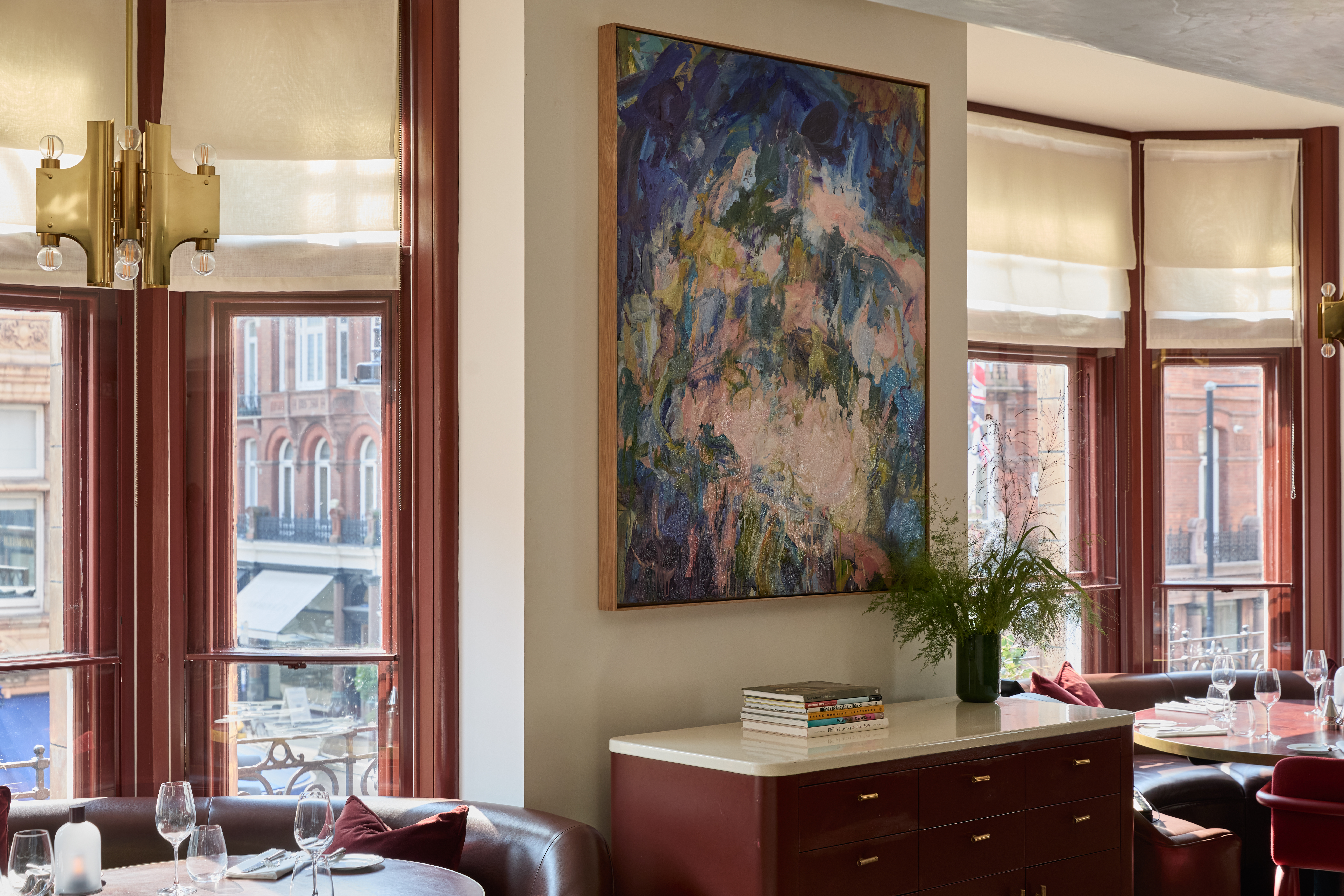 Art restaurants in London - A dining space in Mount St. Restaurant featuring a large abstract painting by Catherine Goodman above a dark red sideboard with books and a vase.