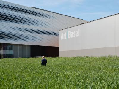 Art Basel - a man walking in grass towards a building with a a wall beside him that says Art Bast