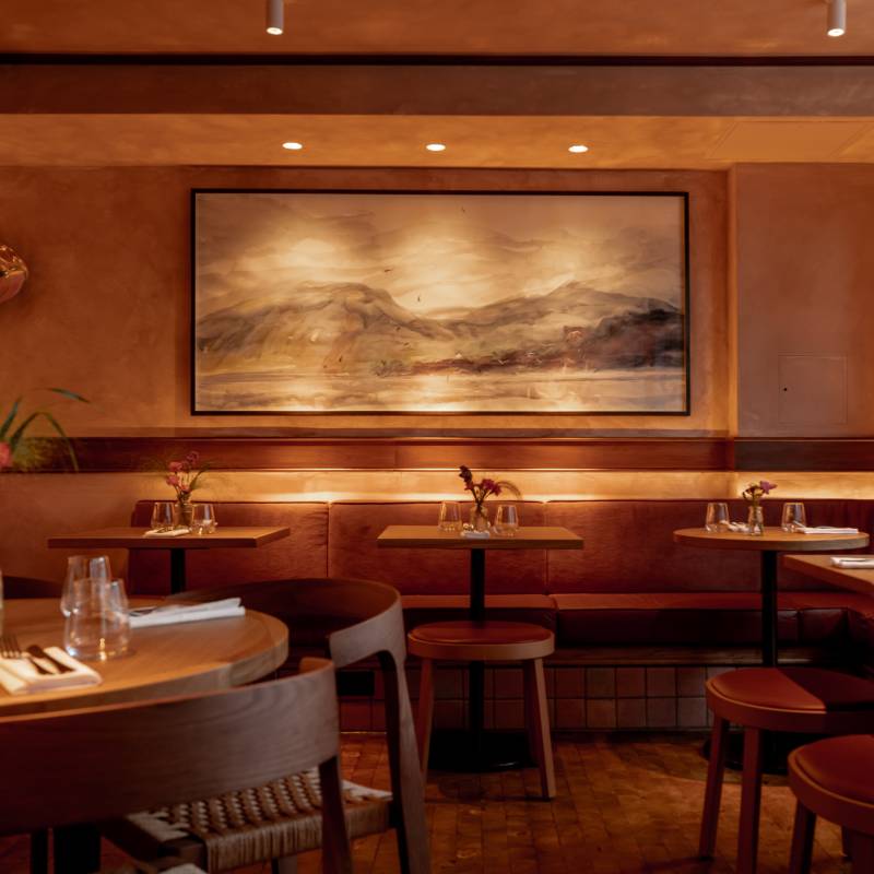 Art restaurants in London - warmly lit restaurant interior of Lolo in Bermondsey featuring wooden tables and cushioned bench seating. A large framed landscape painting serves as the focal point.